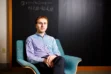 A photo of Harvard mathematician Aaron Landesman sitting in a chair in front of a blackboard.
