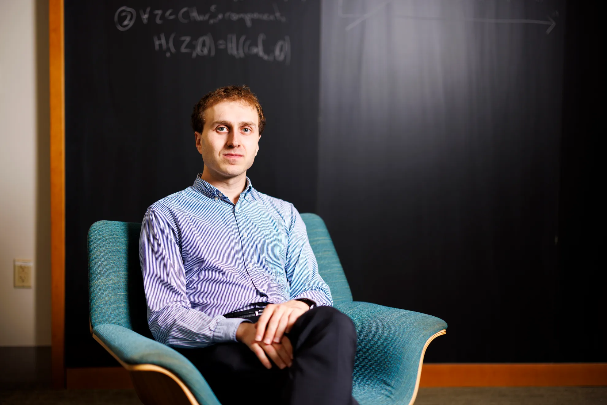 A photo of Harvard mathematician Aaron Landesman sitting in a chair in front of a blackboard.