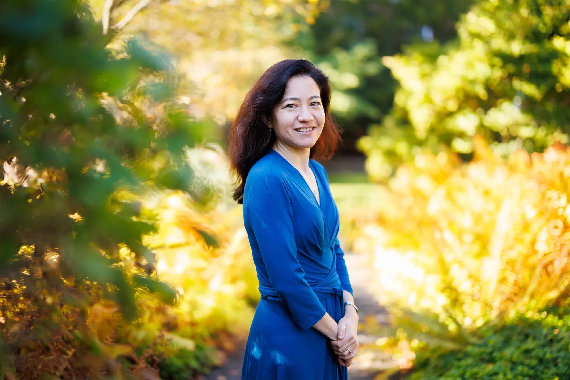 A photo of a woman with long dark hair and a blue dress on the background of blurred foliage.