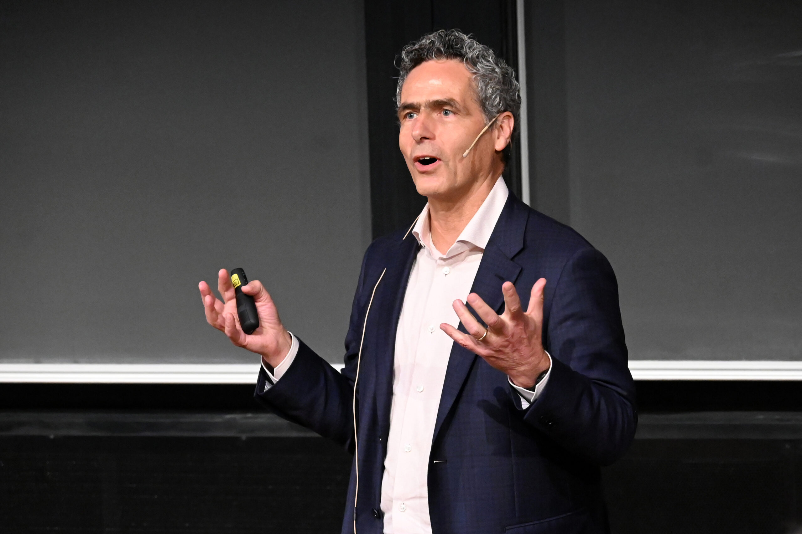 A man wearing a dark blue blazer and a light pink shirt holding a clicker and giving a lecture in front of a blackboard.