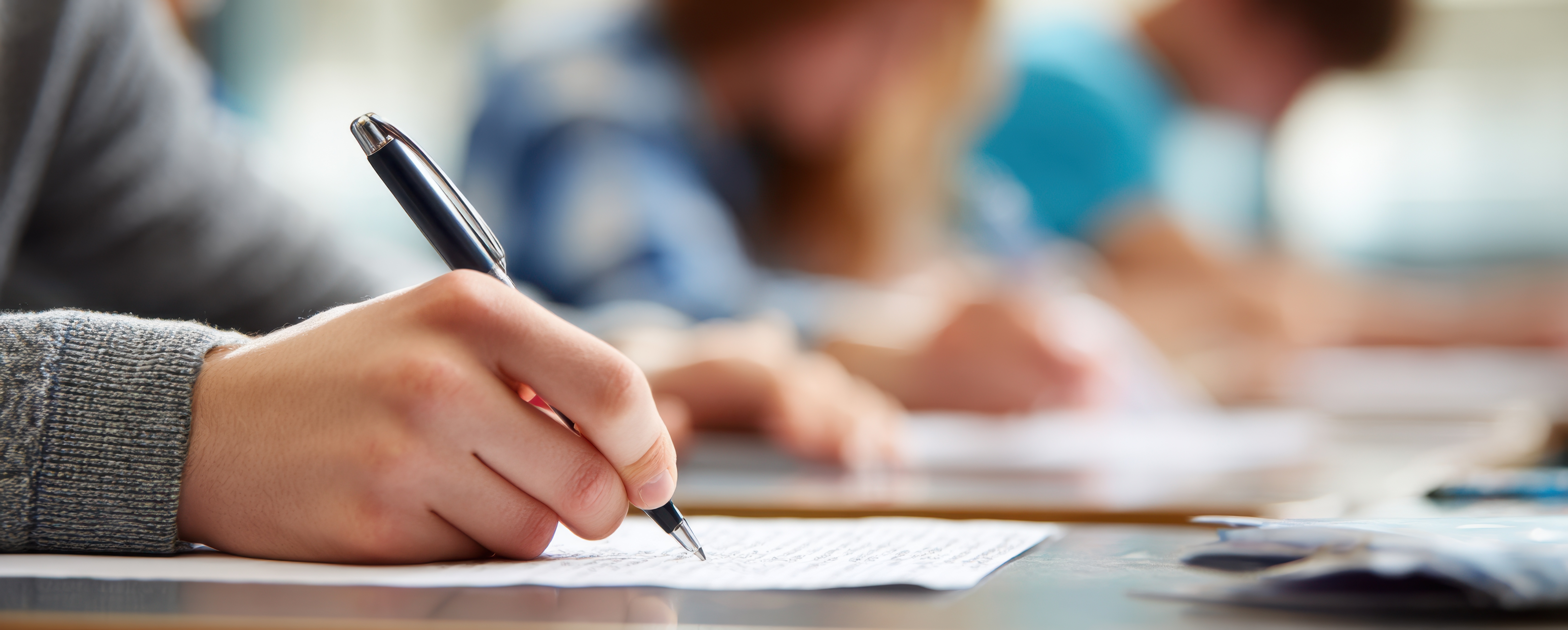 A student is carefully writing on a piece of paper during an exam in a classroom setting.