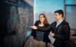 A man and a woman standing in front of a blackboard covered with math formulas as the man gestures at something on the board.