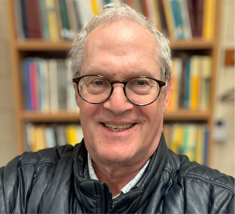 A photo of a man in glasses and a black leather jacket smiling and looking at the camera on the background of a bookcase full of books.