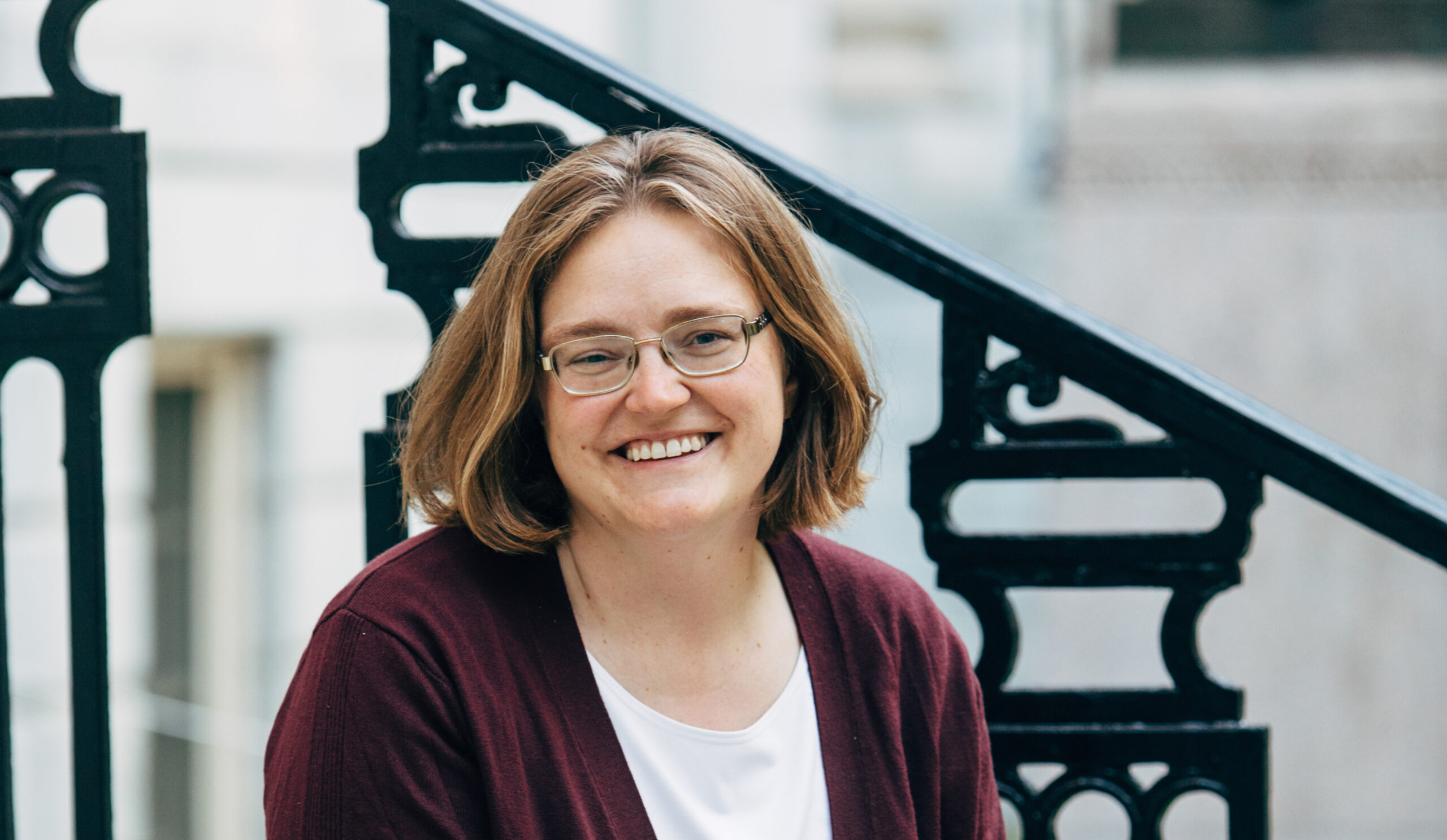 A woman with short blond hair and glasses smiling at the camera on a background of a black wrought iron railing and grey stone.