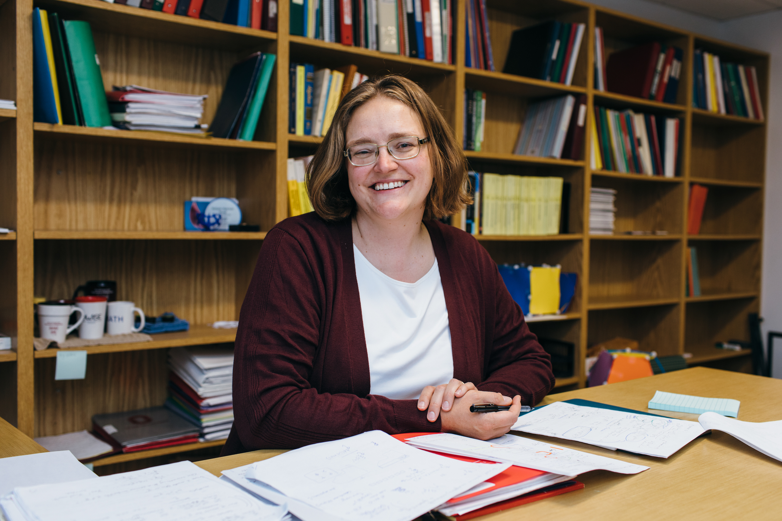 A woman with shoulder-length blond hair wearing glasses and a burgundy cardigan over a white shift is sitting at a desk, holding a pen and smiling at the camera. Behind her is a wall of bookshelves with academic journals, textbooks, and trinkets.