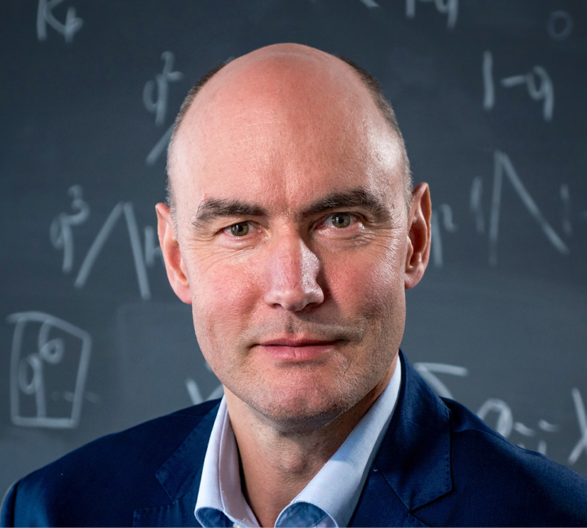 A headshot of a man wearing a navy jacket and light blue shirt looking at the camera. Behind him is a background of math formulas in chalk on a blackboard.
