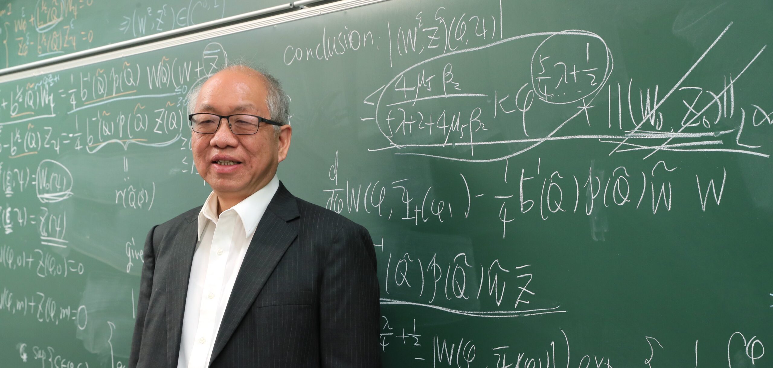 Harvard math Professor Emeritus Shing-Tung Yau against a green blackboard covered in mathematical formulas.
