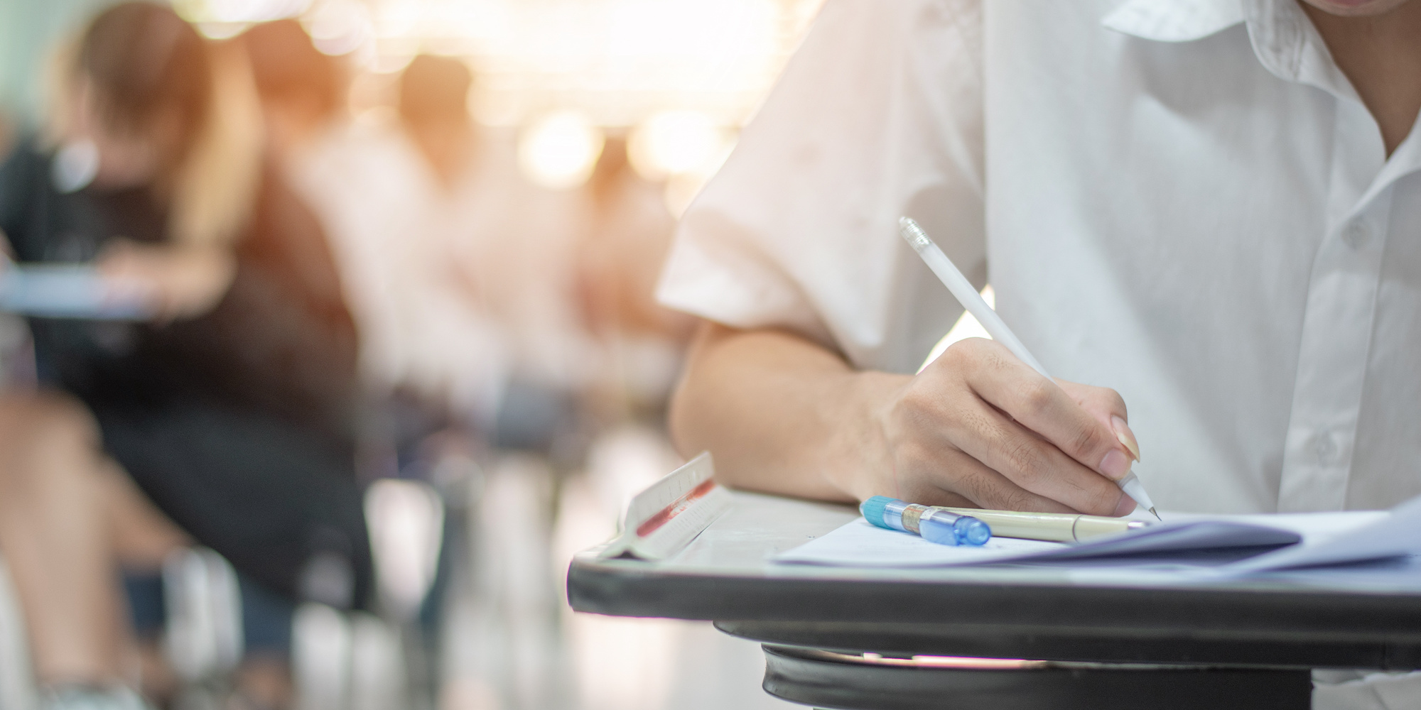 A closeup of a student taking an exam with a row of other students taking an exam out of focus in the background.