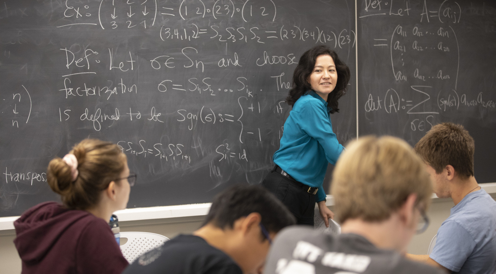 Harvard math professor Lauren Williams teaching a class and writing math formulas in chalk on a blackboard.