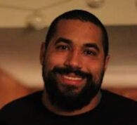 A man with a short beard and dark hair wearing a black sweater is smiling while looking at the camera with a slight head tilt.