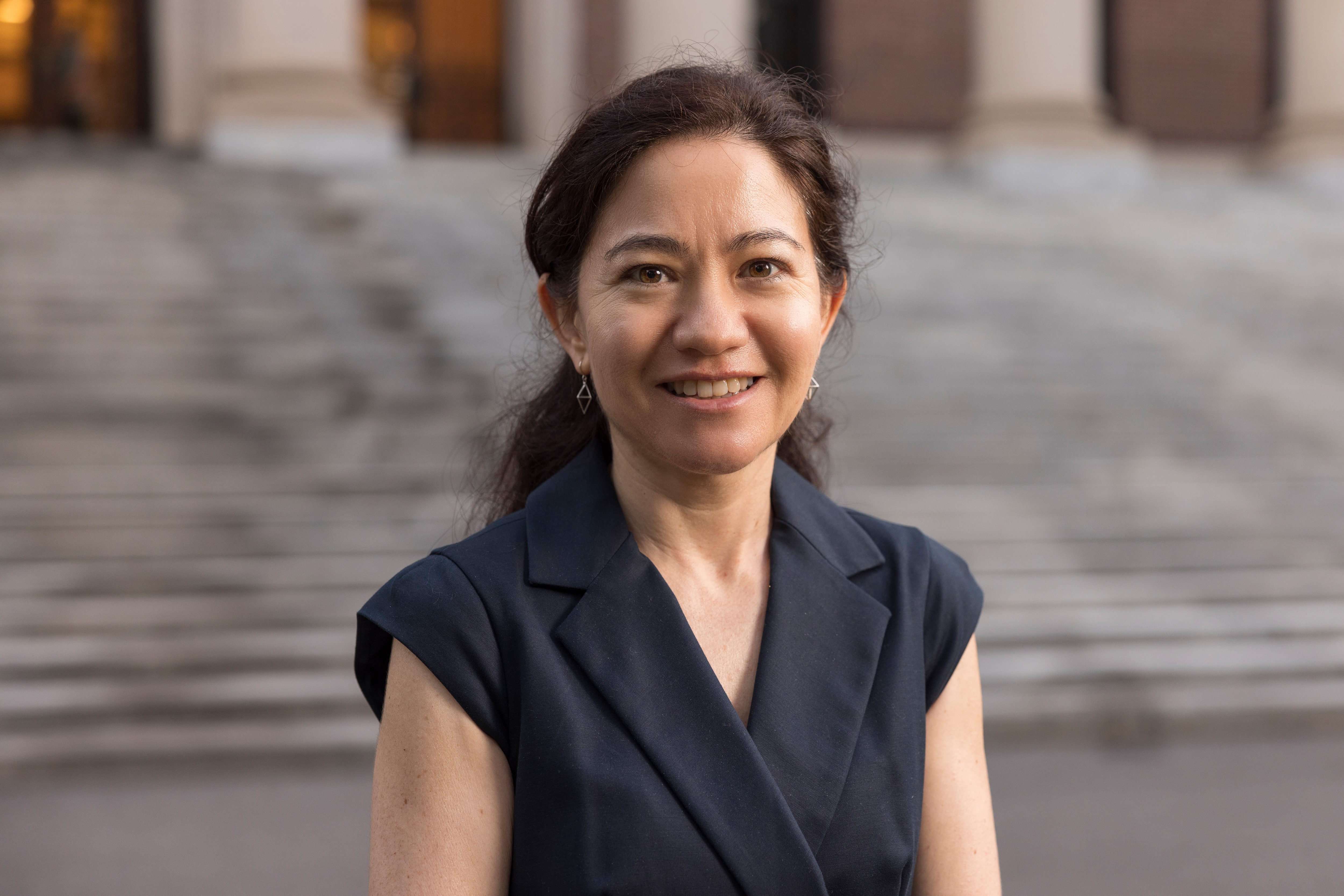 A chest-up photo of Harvard math professor Lauren Williams standing in front of building stairs wearing a sleeveless navy top.